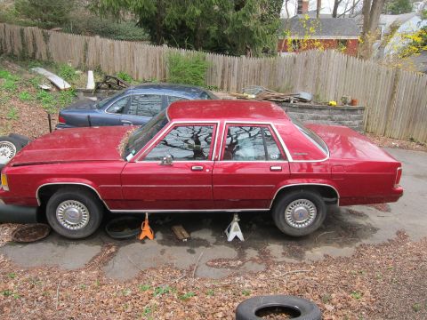 1991 Ford LTD Crown Victoria - A red sedan is raised on jack stands in a driveway, with a blue sedan parked behind it.
