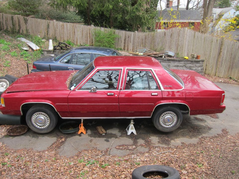 Red sedan on jack stands, another blue sedan behind it. Yard debris and fence in background
