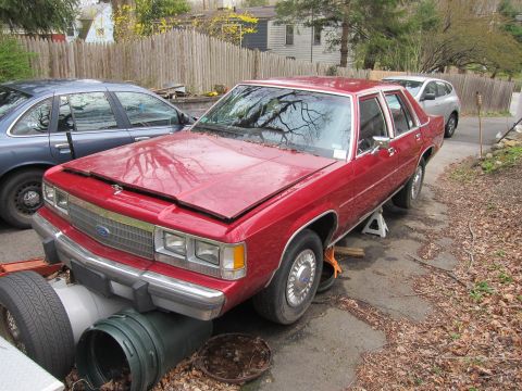 1991 Ford LTD Crown Victoria - A red sedan is elevated on jack stands in a driveway, with another vehicle partially visible behind it.