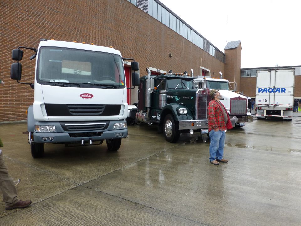 Three trucks parked in a row, a white modern truck in front, a green classic truck in the middle, and a white PACCAR trailer truck in the back