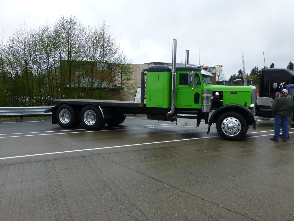 Green flatbed semi-truck parked on wet asphalt, with another black truck partially visible to the right