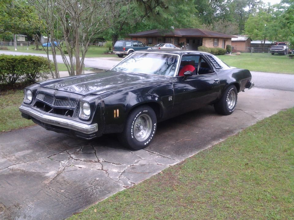 Black coupe parked on a cracked driveway. It has a prominent grille and louvers on the rear side windows