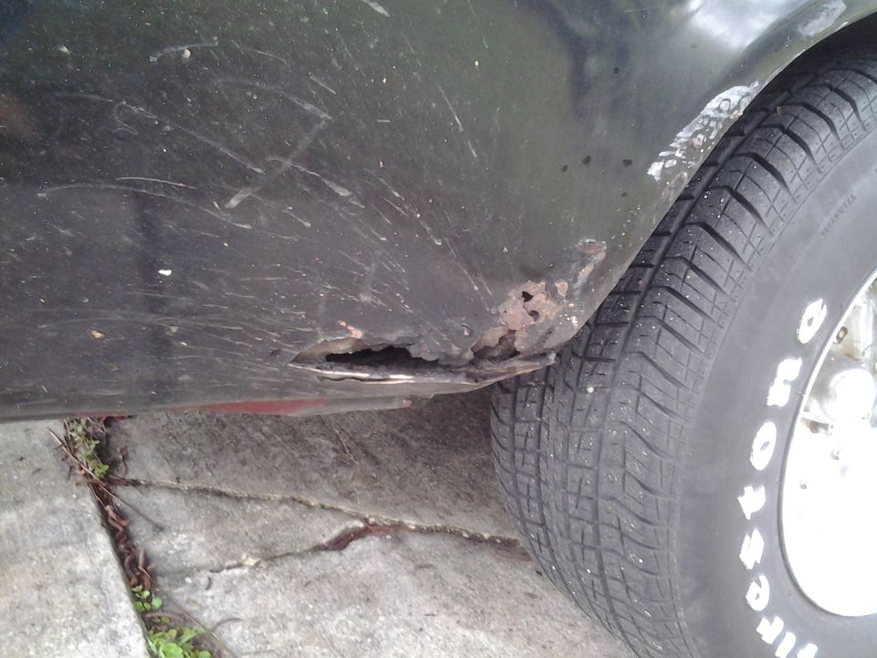 Close-up of a dark green car's rear fender showing significant rust damage and a tire with white lettering