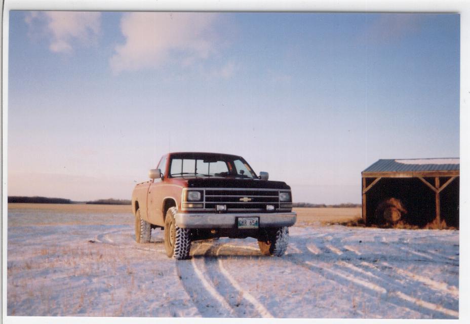 Red lifted Chevrolet square-body pickup in a snowy field beside a shed