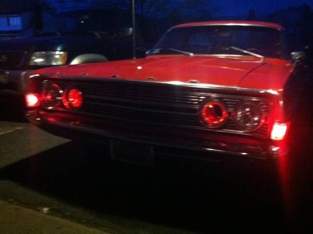 Red 1968 Ford Ranchero front grille and headlights at night, with another vehicle behind it