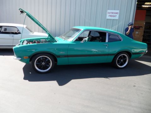 1973 Ford Ranchero - A teal 1970 Ford Maverick coupe is parked outside a building with its hood open, next to a white car.