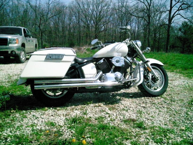 white cruiser motorcycle with saddlebags parked on gravel, silver pickup truck in background