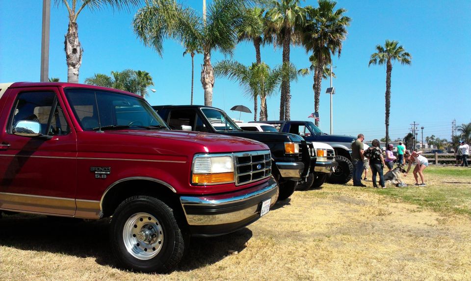 Red 1994 Ford Bronco in front of other trucks and Broncos, parked outdoors with palm trees