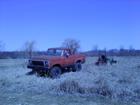 1978 Ford Bronco - A red pickup truck is parked in a field, tethered to a green tractor.