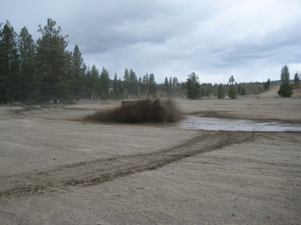 Mud spray erupting from a dirt track in an open field with trees in the background