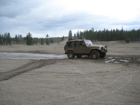 2005 Jeep Wrangler - A Jeep Wrangler navigates a muddy dirt track under an overcast sky, with a dense forest lining the horizon.