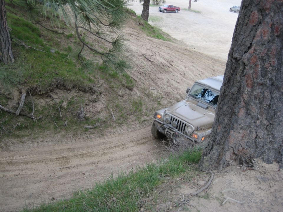 Tan Jeep Wrangler driving on a dirt trail next to a grassy hill. Another red truck and a car are visible in the background