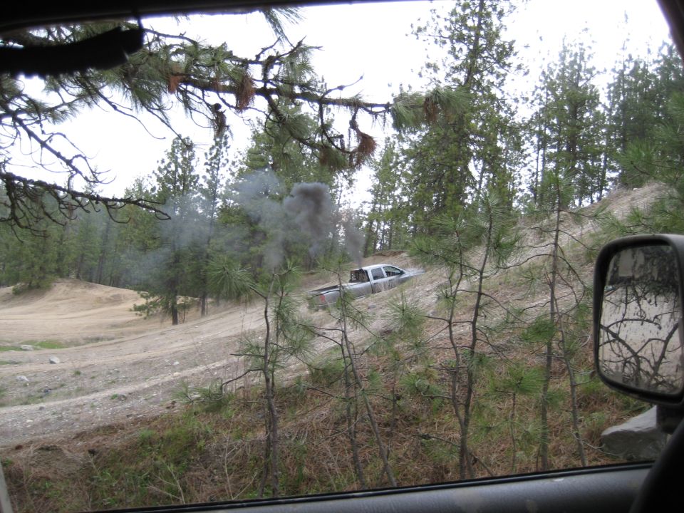Silver pickup truck stuck on a dirt hill, emitting black smoke. Trees and brush surround the scene