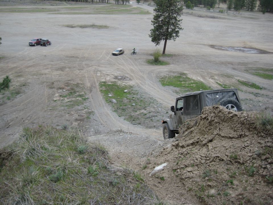 Dark green 2005 Jeep Wrangler driving on a dirt hill, other vehicles and a person on a bike in the background