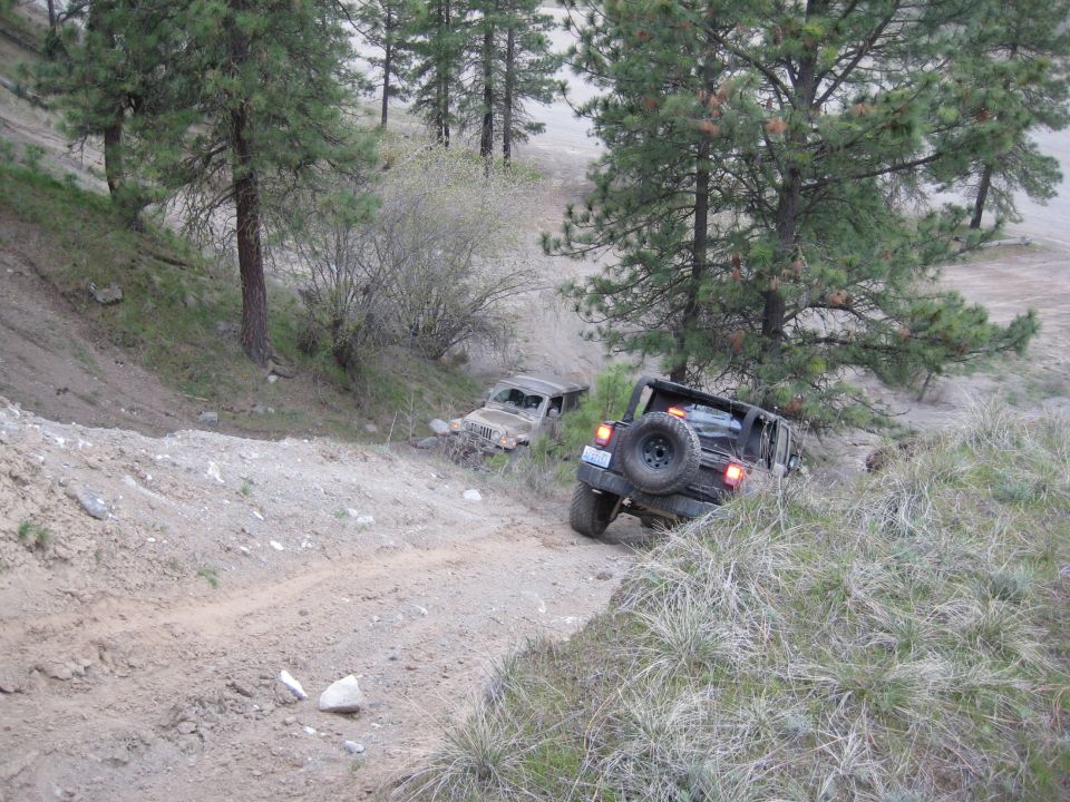 Two Jeep Wranglers driving on a dirt trail in a wooded area. The front Jeep is dark colored, the rear one is lighter