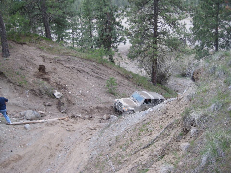 Muddy SUV stuck on a steep, dirt trail with trees and brush around. Person standing on the left