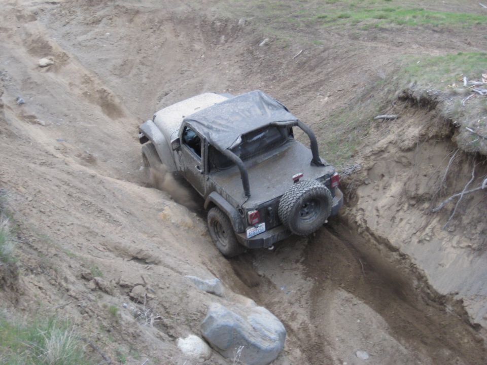 Dark gray Jeep Wrangler with a soft top, angled on a dirt trail, with a spare tire on the rear