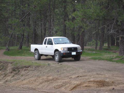 2005 Jeep Wrangler - White Toyota Tacoma pickup truck drives on a dirt road through a wooded area.