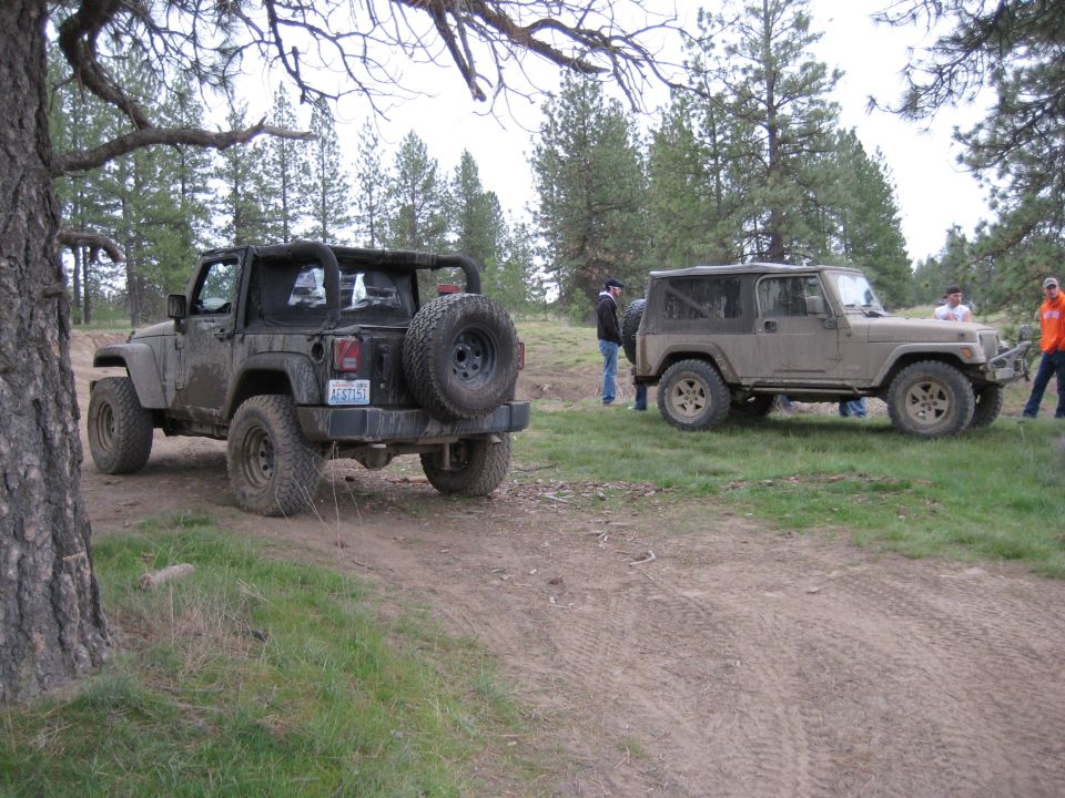 Two Jeep Wranglers, one dark and one tan, parked on a dirt road. The dark Jeep is in the foreground, rear-facing. The tan Jeep is in the background, side-facing