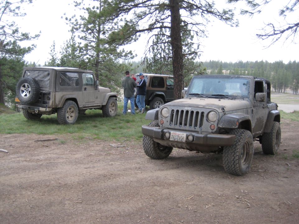 Front view of a dusty Jeep Wrangler parked on dirt, with two other Wranglers and people in the background