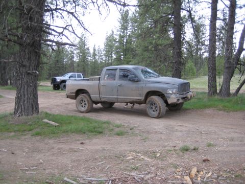 2005 Jeep Wrangler - A gray Dodge Ram pickup truck is parked on a dirt road in a wooded area, with another white pickup truck visible behind it.