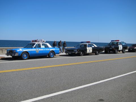 1991 Ford LTD Crown Victoria - Several police cars, including a blue and white model with 'HWY. PATROL' text, are lined up along a coastal road under a clear blue sky.