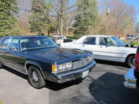 1991 Ford LTD Crown Victoria - Two Ford Crown Victorias are parked outside, one dark blue and the other white with its hood open.