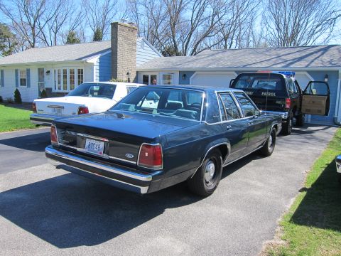 1991 Ford LTD Crown Victoria - A dark blue sedan is parked in a driveway next to a black SUV with 'HIGHWAY PATROL' visible on its side.