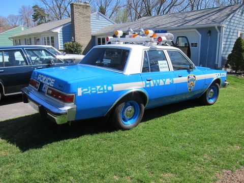 1991 Ford LTD Crown Victoria - A blue and white police car is parked on a grassy lawn in front of a house.