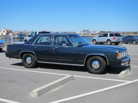 1991 Ford LTD Crown Victoria - A dark blue sedan with chrome accents is parked in a lot near a body of water.