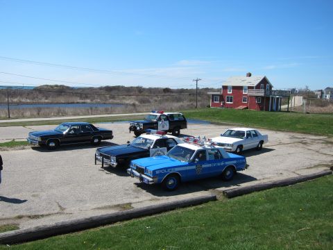 1991 Ford LTD Crown Victoria - A collection of police vehicles and a civilian car are parked outdoors on a sunny day, with a red building in the background.