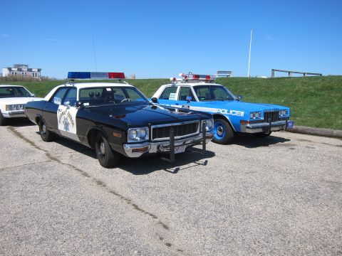 1991 Ford LTD Crown Victoria - Two vintage police cars, one black and one blue, are parked outdoors under a clear sky.