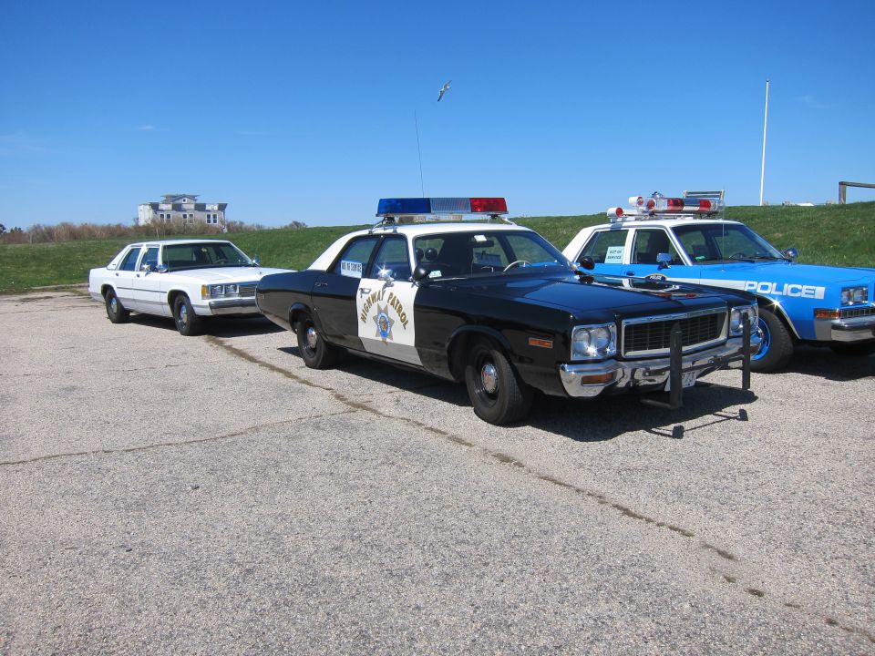 Three police cars parked on asphalt, two with light bars. The front car is black and white with 'HIGHWAY PATROL' on the door. A white car is behind it