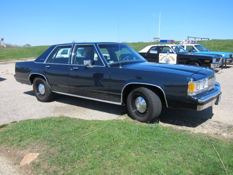 1991 Ford LTD Crown Victoria - A dark blue sedan is parked in front of two police vehicles under a clear blue sky.