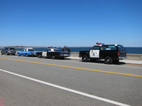1991 Ford LTD Crown Victoria - Several police vehicles, including a 1991 Ford Crown Victoria LTD, are parked along a coastal road under a clear blue sky.