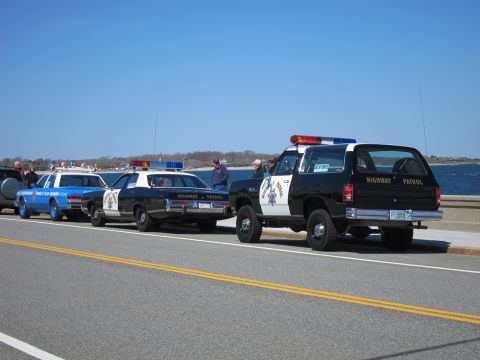 1991 Ford LTD Crown Victoria - Multiple police vehicles are lined up on the side of a road with a body of water in the background.