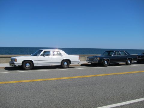 1991 Ford LTD Crown Victoria - Two older model sedans are parked along a road with the ocean in the background.
