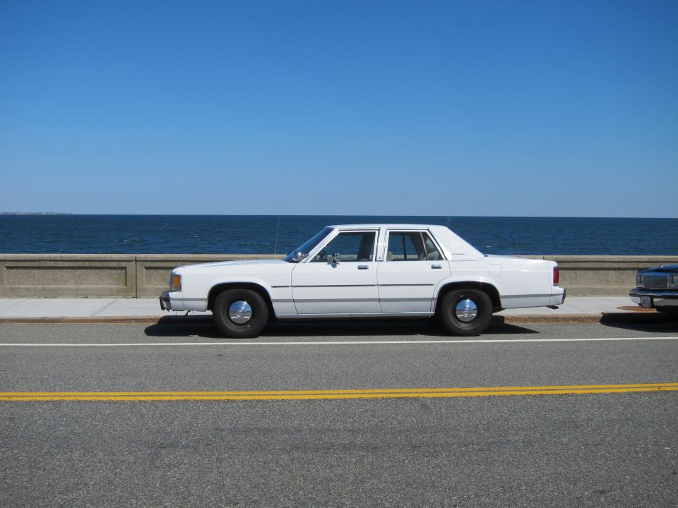White sedan parked by the ocean, side view. It has chrome hubcaps and a clean body