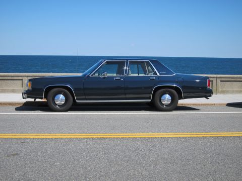1991 Ford LTD Crown Victoria - A dark blue sedan is parked on a road next to a concrete barrier and the ocean under a clear blue sky.