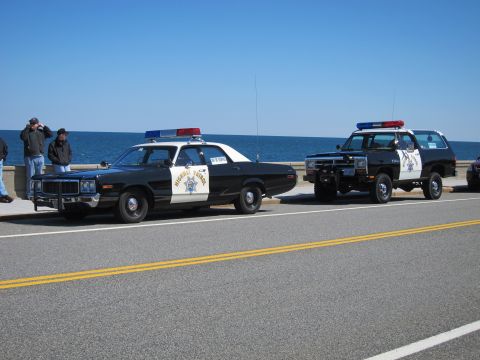 1991 Ford LTD Crown Victoria - Two vintage police vehicles, a sedan and an SUV, are parked side-by-side near the coast under a clear blue sky.