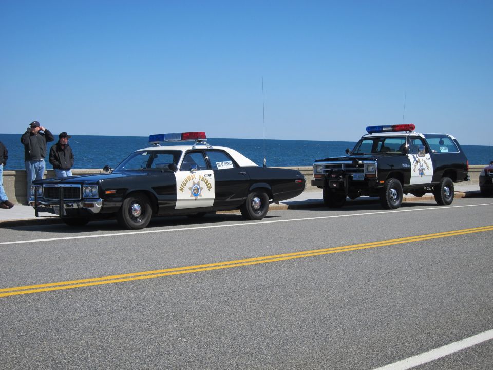 Two police cars, a sedan and an SUV, parked by the ocean. People stand on a wall nearby