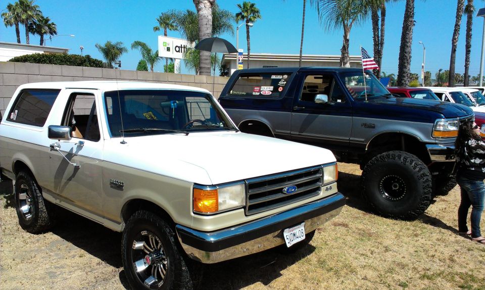 Two Ford Broncos parked outdoors, one white and tan, the other dark blue and gray with large tires