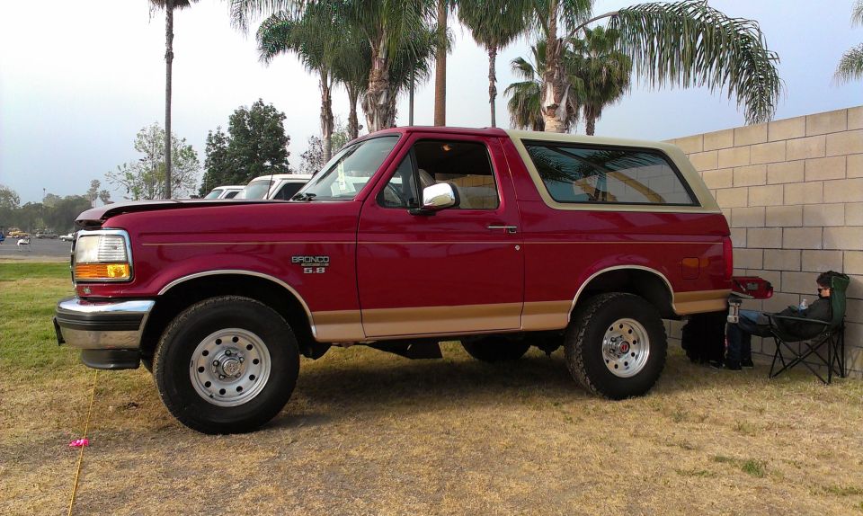 Red Ford Bronco with tan top parked on grass, hood slightly ajar, side view