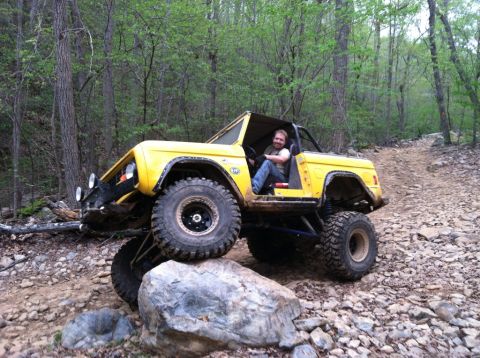 1990 Ford Bronco - This heavily modified yellow Ford Bronco navigates a challenging rocky path through a wooded area.