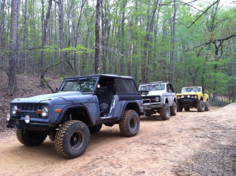 1990 Ford Bronco - Three Ford Broncos driving on a dirt path through a forest with trees on either side.
