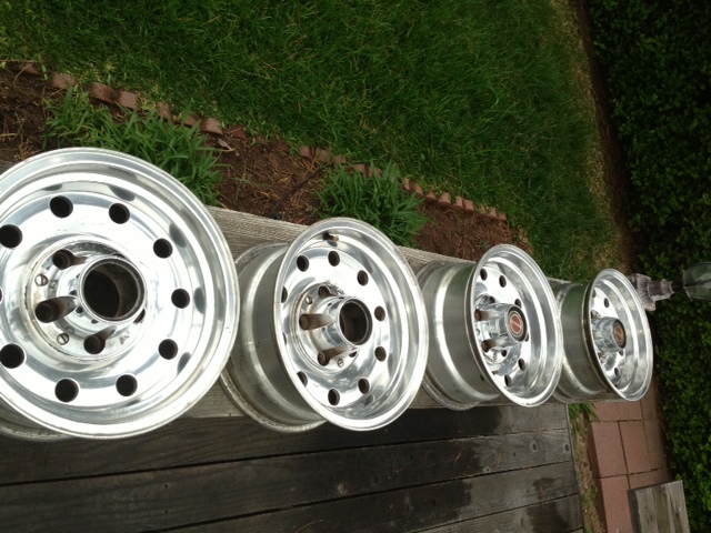 Four aluminum wheels with multiple lug holes and center caps are lined up on a wooden surface