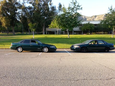 1995 Ford Taurus - Two 1995 Ford Taurus sedans are parked next to each other in a parking lot with trees and a building in the background.