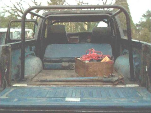 1978 Ford Bronco - The interior of a truck bed is visible, a roll cage, a toolbox with red cables, and a spare tire.
