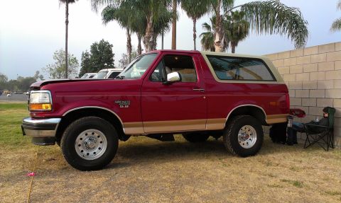 1994 Ford Bronco - A red 1994 Ford Bronco with tan trim is parked on a grassy area next to a brick wall.