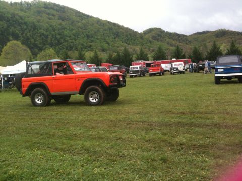 1992 Ford Bronco - A classic orange Ford Bronco is parked on a grassy field, with other vehicles and tents visible in the distance.